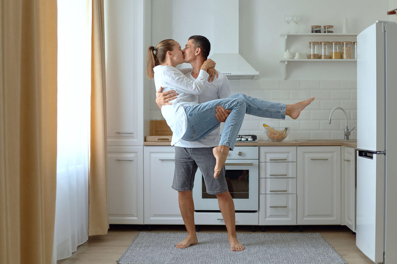 Full length portrait of young man holding his beautiful girlfriend in arms while standing in kitchen at home and kissing her. Both are looking at each other, with love and romantic feelings.